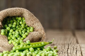 Green peas in open pods, peeled green peas in a burlap bag, scattered pea grains on a wooden background.