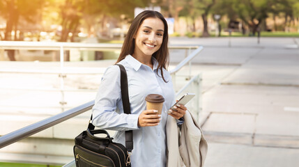 A smiling young woman in business casual attire holding a coffee cup and smartphone stands outdoors