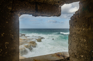 Rocky shore against a cloudy sky and waves in the sea in Malta. Photo taken through a window.