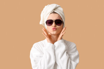 Beautiful woman in sunglasses blowing kiss after shower on beige background, closeup