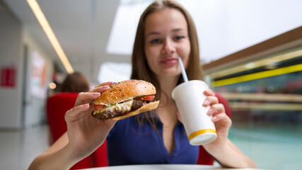 Portrait of young happy woman or beautiful teenager girl eating fast junk food, tasty burger and drink soda in a restaurant or cafe, enjoy meal. 