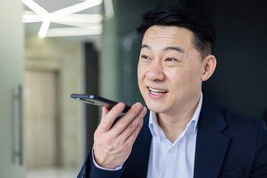 Close-up photo of a smiling Asian businessman sitting in the office, holding the phone and talking on the loudspeaker.