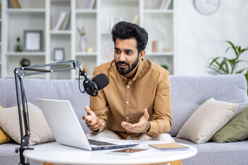 A young Indian man sits at home on the sofa in front of a laptop and speaks into a microphone,...