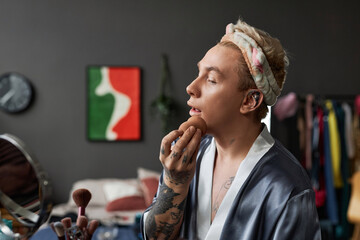 Side view portrait of tattooed young man doing makeup at home and looking in mirror, copy space