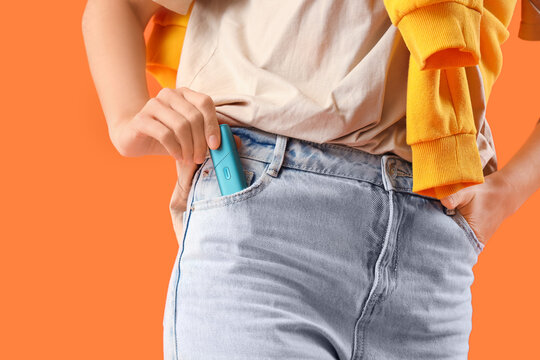 Young woman with electronic cigar in jeans pocket on orange background, closeup