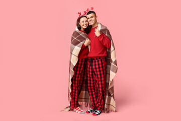Happy young couple in Christmas pajamas and with plaid  hugging against pink background - obrazy, fototapety, plakaty