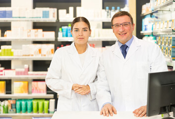 Fototapeta na wymiar Two smiling female and male pharmacist workers stand shoulder to shoulder and affably welcoming visitors in pharmacy s sales area