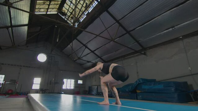 Professional female acrobat with tattoos on muscular body doing cartwheel and backflip on mat while exercising in gym. Slow motion shot