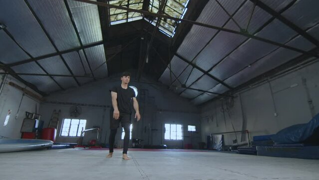 Young male acrobat doing backflip during stunts training session in spacious gym. Full length shot, slow mo