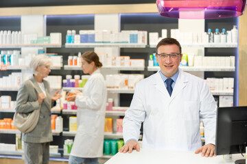 Positive middle-aged male pharmacist posing in chemist's shop with large assortment of goods
