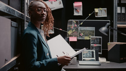 African american agent reviewing evidence to solve crime with clues and criminal records, working in police archive. Woman criminologist standing near desk, looking at detective map board. - obrazy, fototapety, plakaty