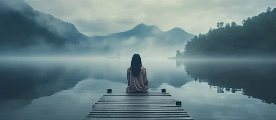 Zelfklevend Fotobehang Back view of fashioned young woman sitting on wooden dock looking at view on a misty morning Female hipster with brown hat relaxes on the edge of jetty admiring foggy lake Wonderful nature geta © Ilgun