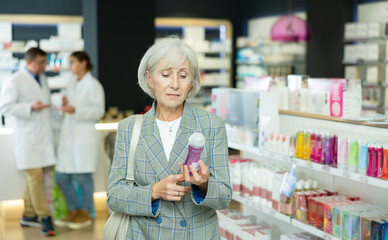 Near self-service window in pharmacy, senior female customer chooses lubricant gel, carefully reads...