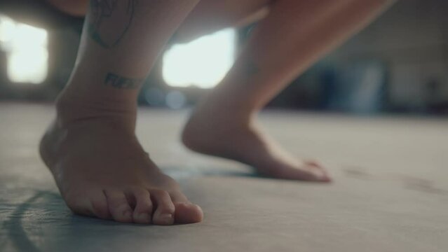 Feet of female gymnast with tattooed leg kneeling and crouching on the floor in gym during training session. Close-up view, ground level shot