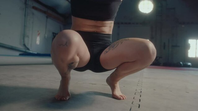 Muscular female gymnast wearing sports bra and short kneeling and crouching in gym while doing balance exercise. Low angle view