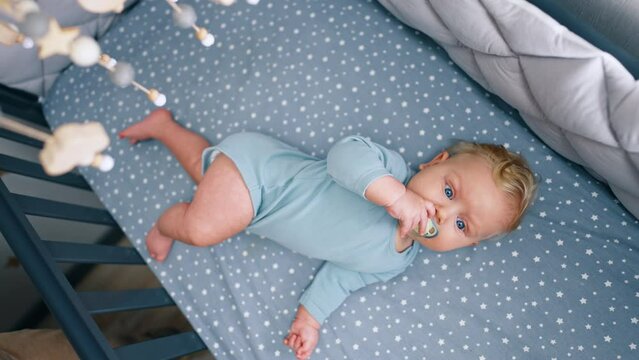 Adorable little baby boy in blue bodysuit lies in the crib. Cute child with pacifier in mouth looks at cot mobile. Top view.