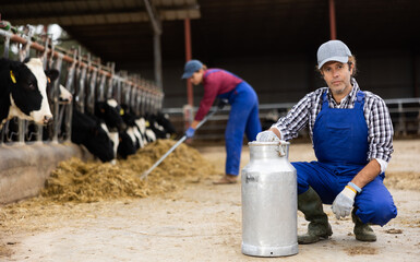Confident hardworking man, owner of farm in overalls with metallic milk can on livestock farm in...