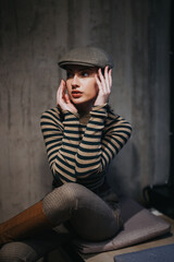 Obraz premium Attractive young woman in her 20s with a cheerful expression, sitting indoors and wearing a stylish newsboy hat. Her striped clothing adds to her fashionable and elegant look.