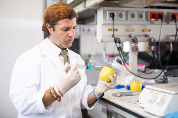 Middle-aged male scientist injecting reagent from syringe into apples, performing scientific...