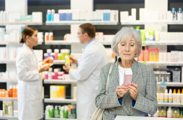 Near self-service window in pharmacy, senior female customer chooses cream for aging skin,...