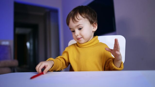 Beautiful Caucasian toddler sitting at desk at home. Lovely baby is focused on drawing with a pen.