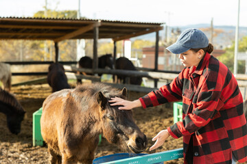 Skilled young female worker in plaid shirt standing close to paddock with foals in countryside horse club