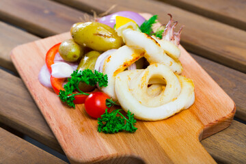 Delicious baked squid rings with potatoes and fresh vegetables and herbs served on wooden board