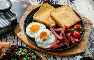 Delicious breakfast with fried eggs, toasts, sausage and vegetables served on hot pan over wooden background. Healthy breakfast, close up