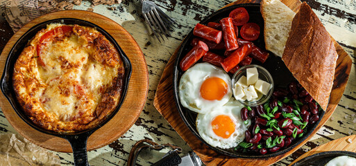 Set of delicious breakfasts, fried eggs with toasts and sausage on hot pan, english breakfast with beans and omelette with cheese on pan over wooden background. Healthy food, top view - obrazy, fototapety, plakaty