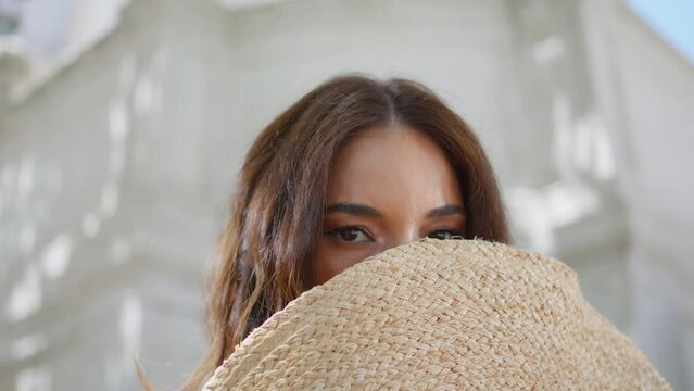 Playful woman hiding face straw hat city portrait. Elegant lady looking camera