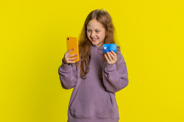Happy smiling preteen child girl kid using credit bank card and smartphone while transferring spending money, purchases online shopping, ordering food delivery. Children isolated on yellow background