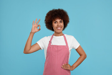 Happy young woman in apron showing ok gesture on light blue background