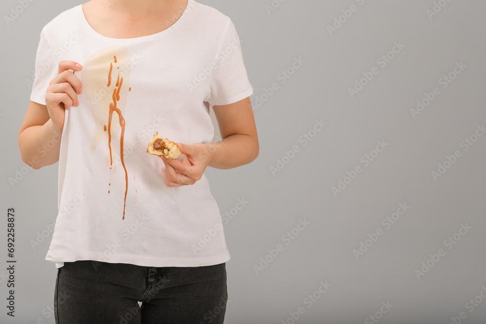 Poster Woman holding pastry and showing stain from condensed milk on her shirt against light grey background, closeup. Space for text