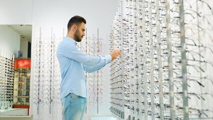 Side view of male customer choosing eyewear while standing near shelf during shopping at optical shop
