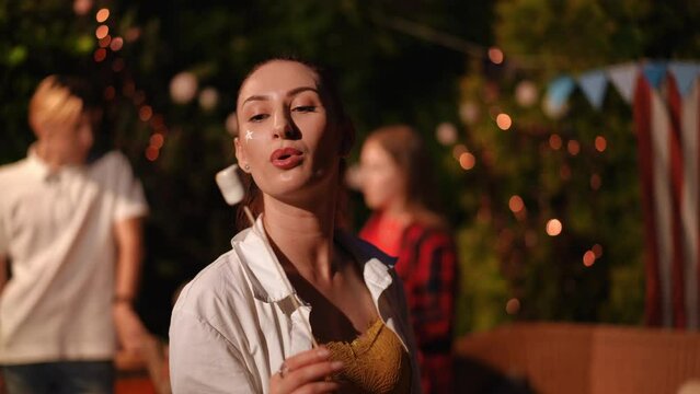 A beautiful girl at a party holds a stick with marshmallows roasted on a fire in her hands and blows on it, looks at the camera and smiles