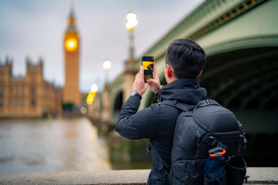 Unrecognizable young Latin man tourist in casual clothes standing alone against ancient cathedral in London near tower bridge and Big Ben against clock tower and taking photos while sightseeing