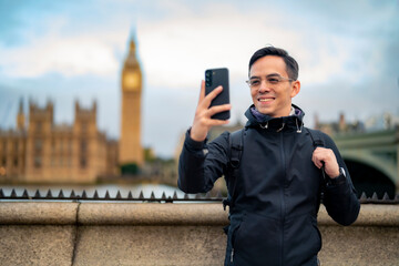 Positive young Latin male tourist standing on embankment and using smartphone in front of historic...