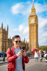 Smiling young Latin male in casual clothes and sunglasses using smartphone while standing on city...