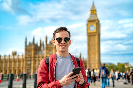 Smiling young Latin male in casual clothes and sunglasses using smartphone while standing on city street during vacation trip to London against Big Ben and clock tower
