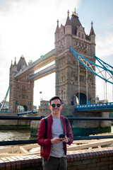 Happy young Latin man wearing sunglasses and red jacket with backpack using smartphone near metal...