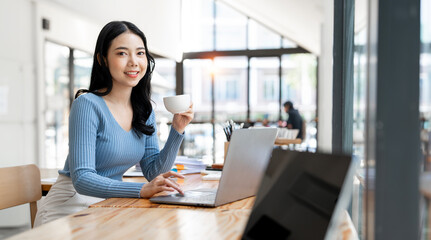 Smiling woman holding coffee cup and using laptop while sitting at co-workspace.