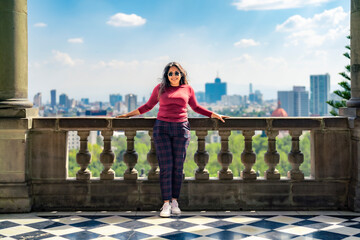 Full body of stylish young Latin woman tourist in casual clothes and sunglasses standing near stone railing of Chapultepec Castle in Mexico city against blurred cityscape background of Reforma street - Powered by Adobe