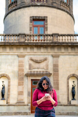 Fototapeta na wymiar Happy young smiling Latin woman tourist in casual clothes and sunglasses using smartphone while standing against blurred historic Chapultepec Castle in Mexico City during trip