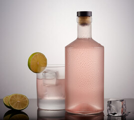 Bottle and glass of pink gin with ice and a lemon slice on a cool gradient background.