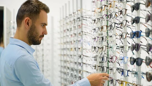 Side view of male customer choosing eyewear while standing near shelf during shopping at optical shop