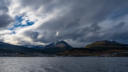 A picturesque snow-capped mountain range of the Andes against a cloudy sky. The town houses of Ushuaia are visible at the foot of the mountains, on the banks of the Beagle Canal. Argentina