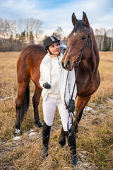 Beautiful blond professional female jockey standing near brown horse in field in winter. Friendship with horse concept