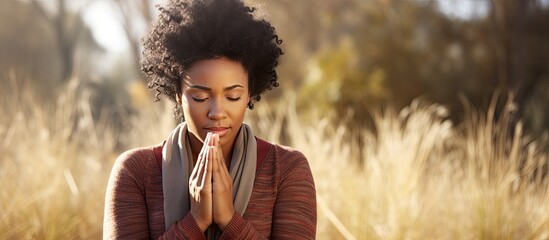 Attractive young African American woman praying in serene park, hands clasped.