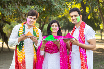 Happy group of three indian people wearing white kurta holding colorful paint or gulal plate in...