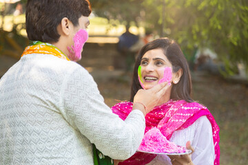 Happy young indian couple wearing white kurta playing, putting colour on face. Celebrating holi together outdoor at park or garden.